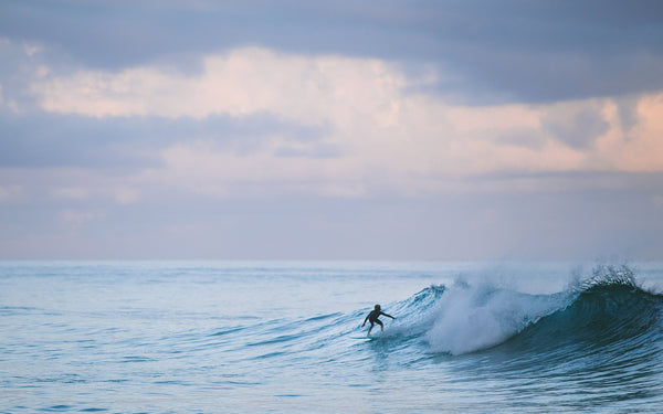 A grommet surfing a clean breaking wave, with peachy clouds in the background.