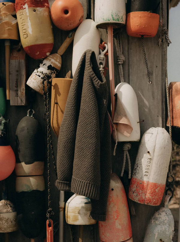 The Fisherman Sweater in Taupe Merino hanging on a wall of buoys