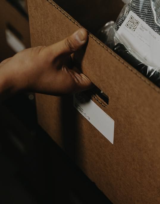 A hand pulls a cardboard drawer from a shelf. It contains a button down shirt in a transparent poly bag. A label bearing the words Taylor Stitch is visible near the collar.