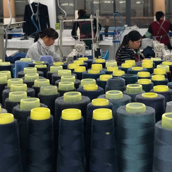 Dozens of spools of dark-colored thread frame a group of four women working at sewing machines in a factory.